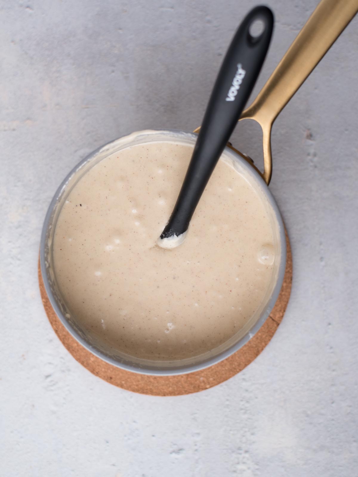 A pot filled with creamy, eggnog fudge sauce sits on a cork mat. A black spoon rests in the speckled, smooth mixture, and the pots handle gleams golden against the light gray background.