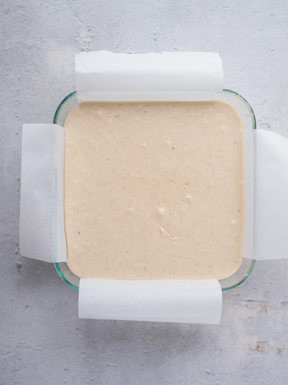 Overhead view of a square glass baking dish lined with parchment paper, filled with light beige eggnog fudge batter, set on a textured gray surface.