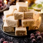 A stack of creamy eggnog fudge squares is arranged on a plate. The background is decorated with festive lights, red ornaments, and sugared cranberries, creating a holiday atmosphere. Text at the bottom reads Eggnog Fudge.