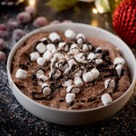 A bowl of chocolate dip topped with mini marshmallows and a drizzle of chocolate sauce. The dessert is set on a dark surface, sprinkled with sugar, and surrounded by festive decorations like red ornaments and sugared berries.