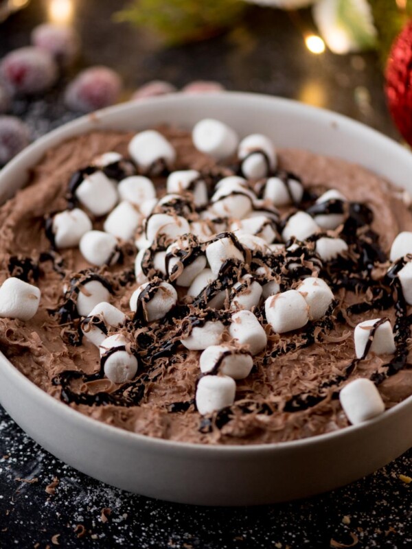 A bowl of chocolate dip topped with mini marshmallows and a drizzle of chocolate sauce. The dessert is set on a dark surface, sprinkled with sugar, and surrounded by festive decorations like red ornaments and sugared berries.