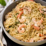 A pan filled with creamy shrimp linguine, featuring perfectly cooked shrimp scattered throughout. The dish is garnished with chopped herbs and appears rich and flavorful. Utensils and a striped napkin are partially visible beside the pan, completing this inviting shrimp pasta scene.