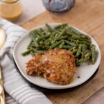 A plate with crispy almond chicken topped with gravy, served alongside green beans with seasoning. The table setting includes a striped cloth napkin and a glass of sauce, all set on a wooden surface.