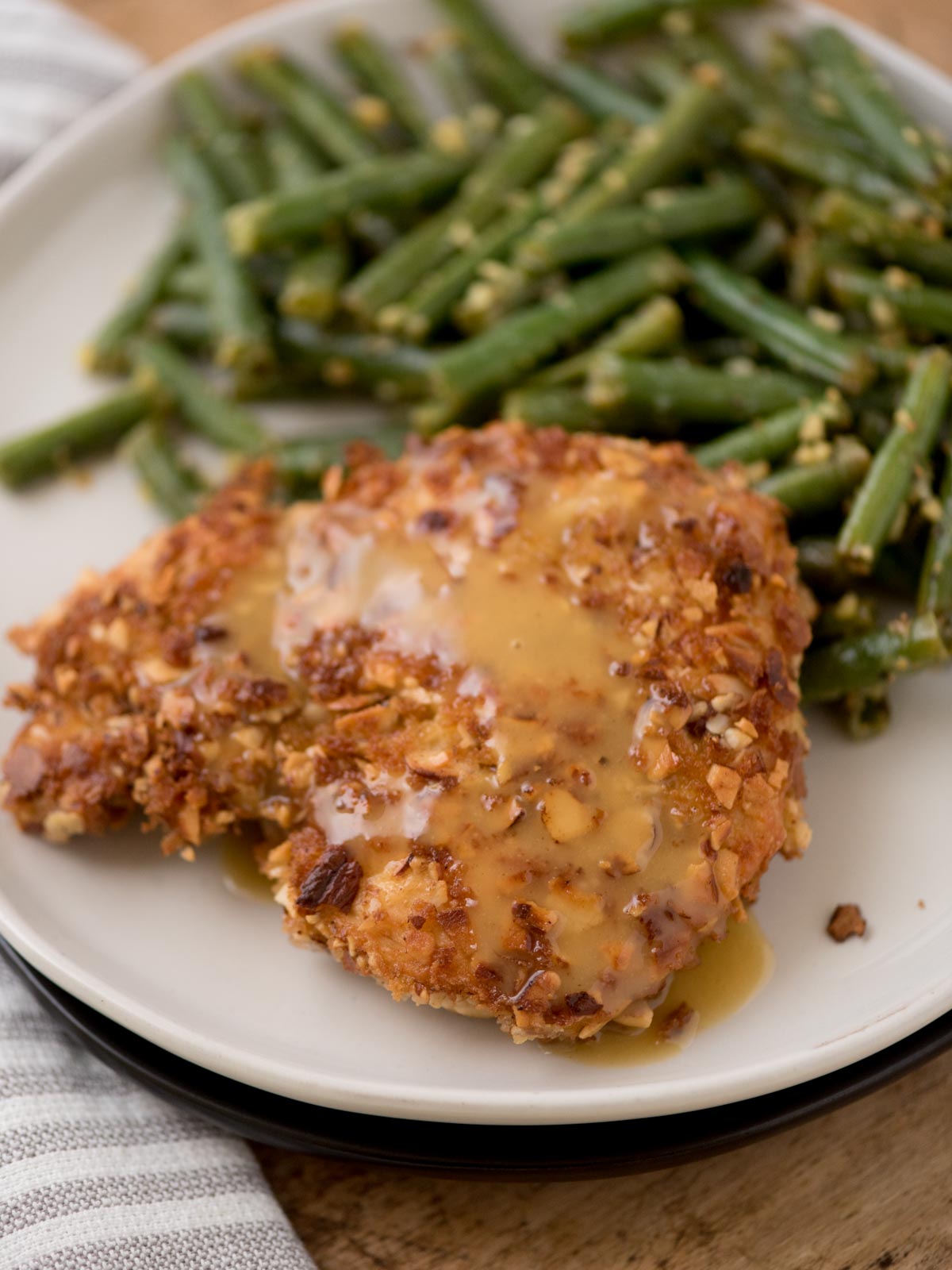 A plate featuring a crispy almond chicken breast topped with a brown sauce, accompanied by green beans seasoned with garlic. The meal is set on a white plate with a gray and white striped napkin beside it.