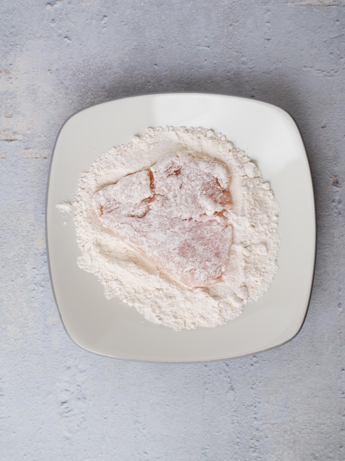 A piece of raw meat, destined to become almond chicken, is coated in flour and rests on a square white plate. The plate sits elegantly on a textured, light gray surface.