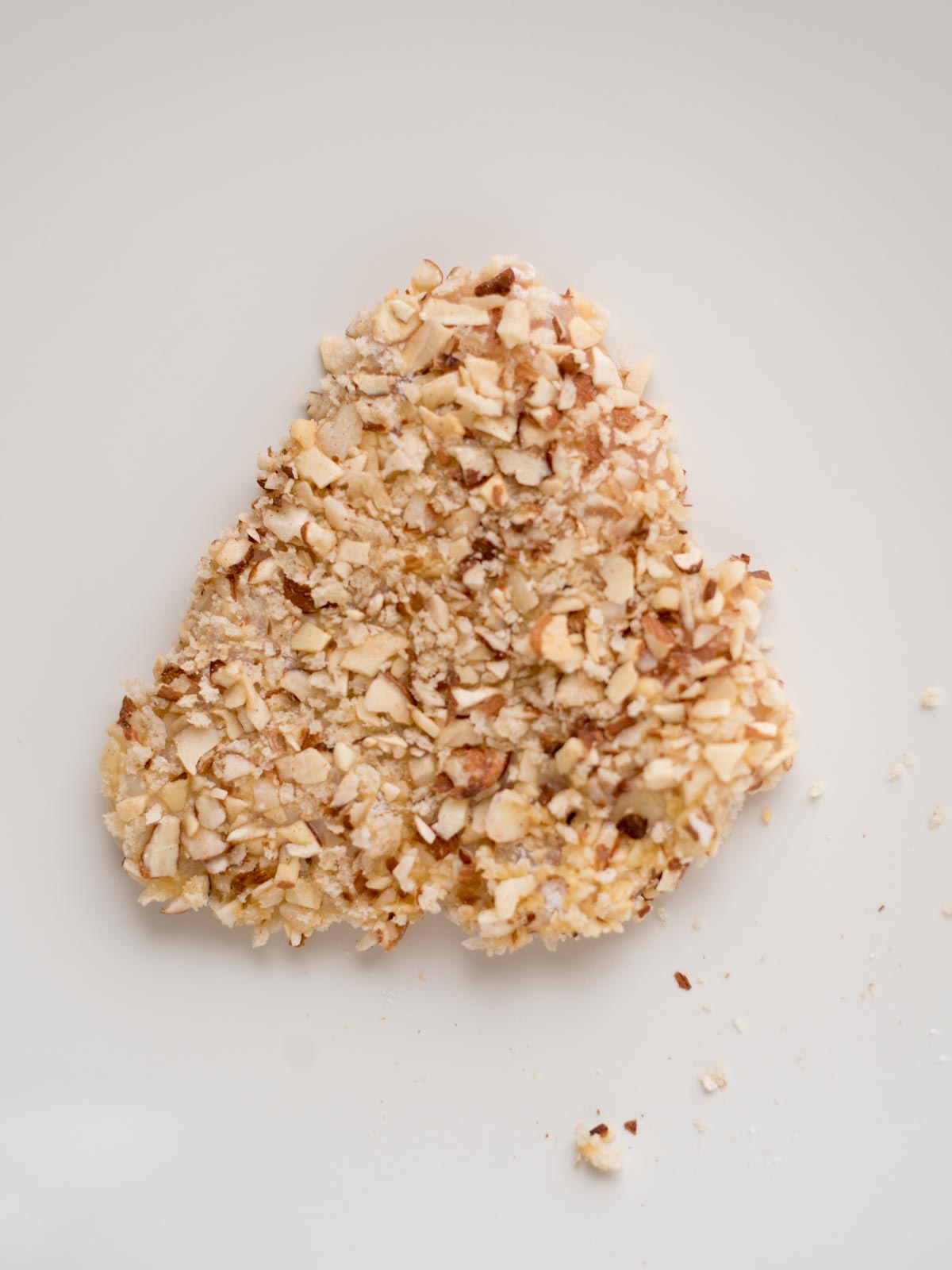 A heart-shaped food item coated with a crunchy almond crust, resting on a plain white surface. Some nut crumbs are scattered around it.