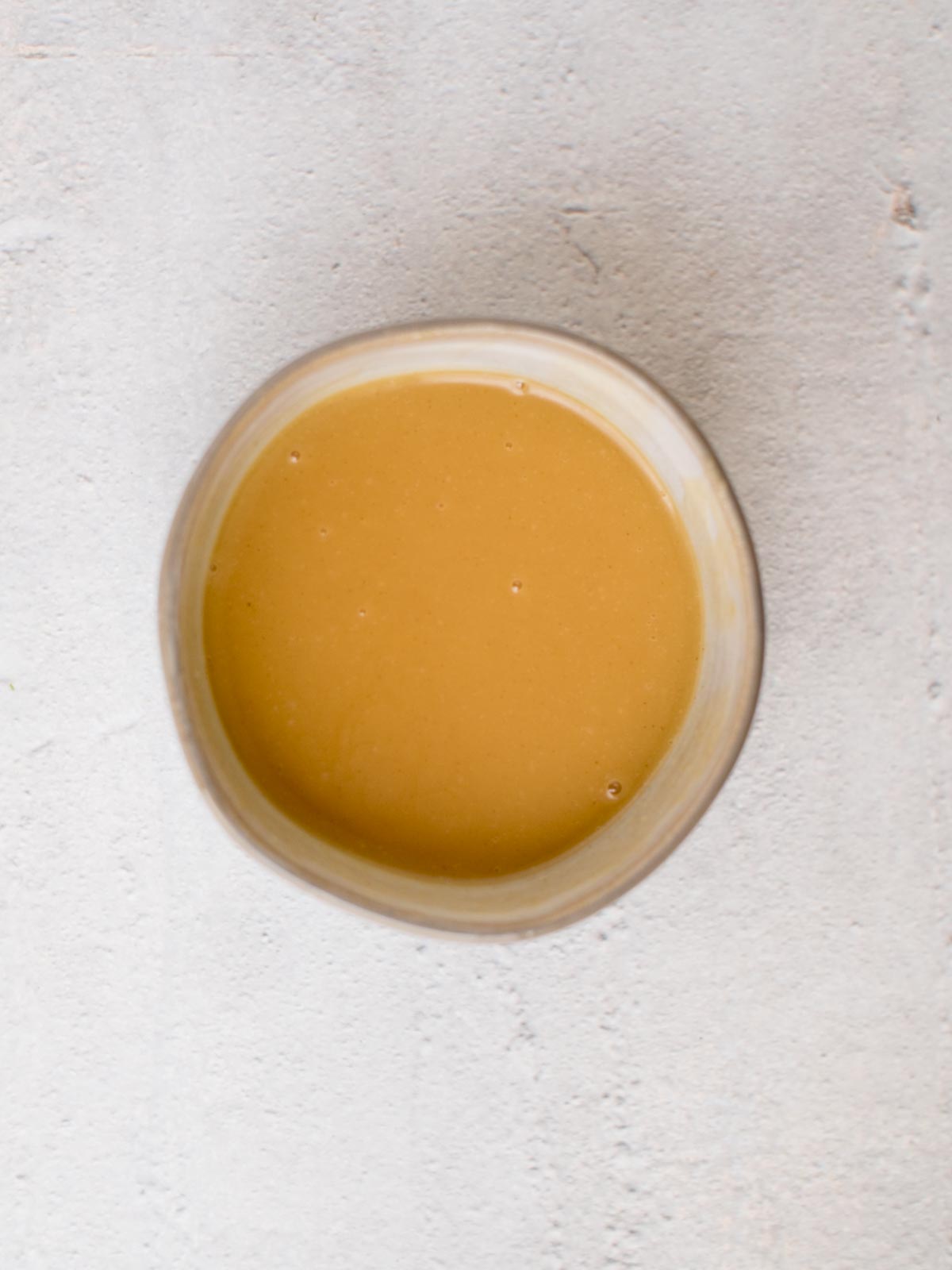 A round bowl filled with smooth, light brown sauce sits on a textured gray surface, perfectly complementing the flavors of crispy almond chicken.