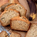 Slices of banana bread on a wooden board, with a partially visible ripe banana in the background. The bread has a golden crust and a moist, speckled interior, and a knife is partially visible beside it.