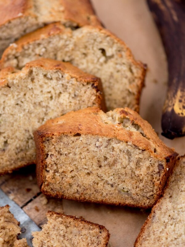 Slices of banana bread on a wooden board, with a partially visible ripe banana in the background. The bread has a golden crust and a moist, speckled interior, and a knife is partially visible beside it.