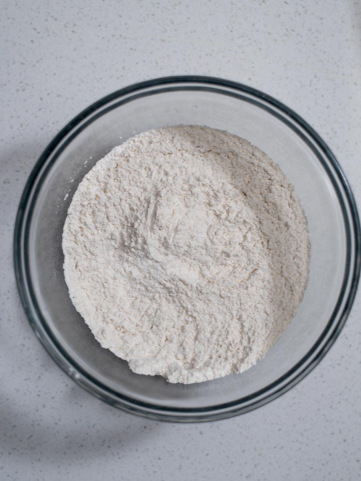A glass bowl filled with white flour sits on a speckled white countertop. The flour appears finely milled and is slightly mounded in the center of the bowl.