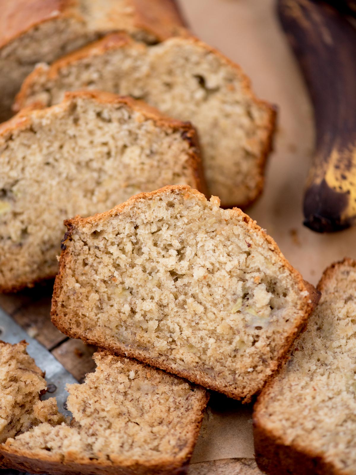 Slices of freshly baked banana bread on a wooden surface, showcasing its moist and fluffy texture. A partially visible ripe banana is in the background, adding context to the flavor of the bread.