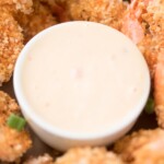 Close-up of a small white bowl filled with creamy dipping sauce, surrounded by golden-brown breaded shrimp. The crispy coating on the shrimp is speckled with herbs. Green onion pieces are scattered among the shrimp.