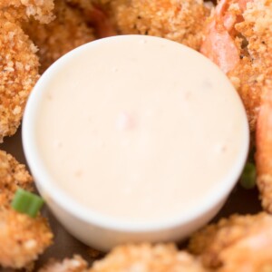 Close-up of a small white bowl filled with creamy dipping sauce, surrounded by golden-brown breaded shrimp. The crispy coating on the shrimp is speckled with herbs. Green onion pieces are scattered among the shrimp.