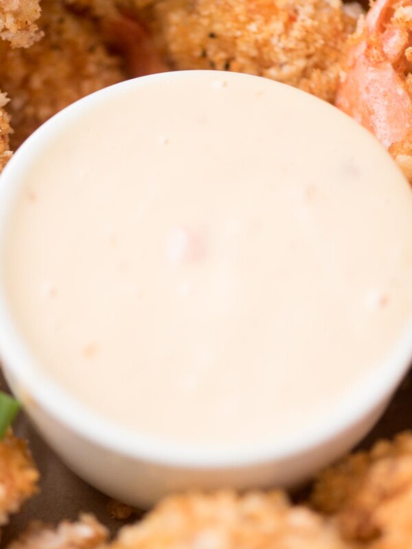 Close-up of a small white bowl filled with creamy dipping sauce, surrounded by golden-brown breaded shrimp. The crispy coating on the shrimp is speckled with herbs. Green onion pieces are scattered among the shrimp.