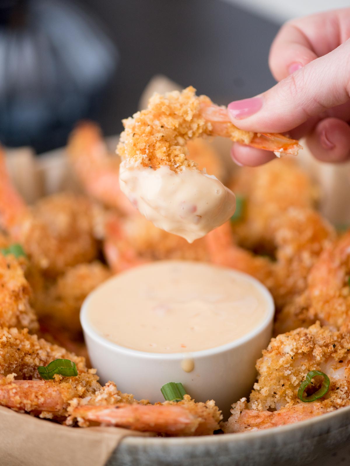 A hand dips a piece of breaded shrimp into a white bowl of creamy sauce. The shrimp are arranged around the bowl on a plate, garnished with green onions.