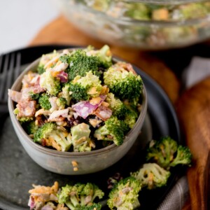 A bowl of broccoli salad with chopped red onions, shredded cheese, and bits of bacon. The salad is in a small grey bowl on a dark plate, with some salad spilled around it. A larger bowl with more salad is blurred in the background.