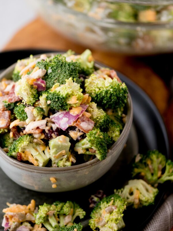 A bowl of broccoli salad with chopped red onions, shredded cheese, and bits of bacon. The salad is in a small grey bowl on a dark plate, with some salad spilled around it. A larger bowl with more salad is blurred in the background.