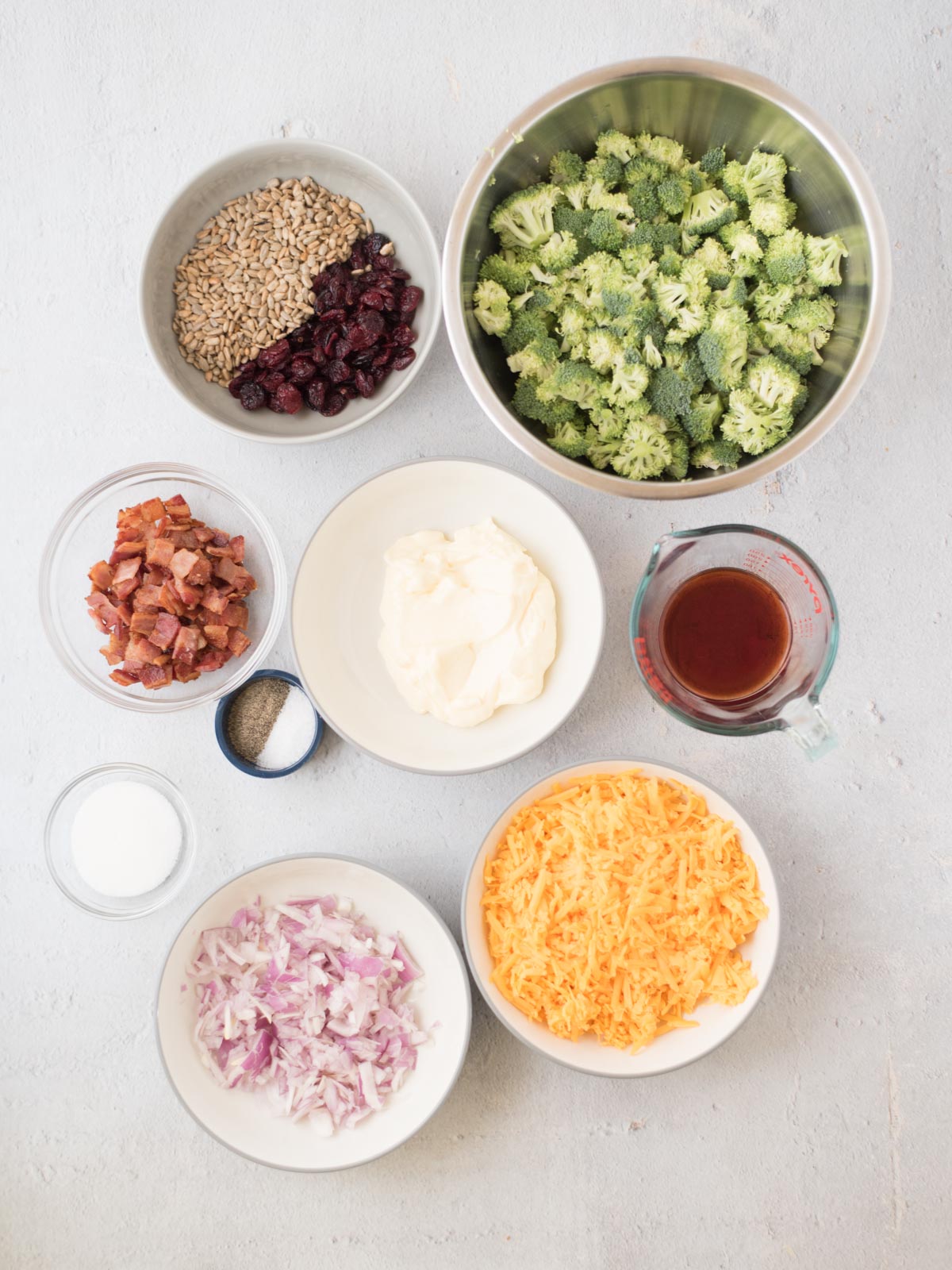 Ingredients on a table include chopped broccoli, shredded cheese, mayonnaise, diced red onion, cooked bacon bits, sunflower seeds, dried cranberries, a small bowl of sugar, and a measuring cup with a dark liquid.