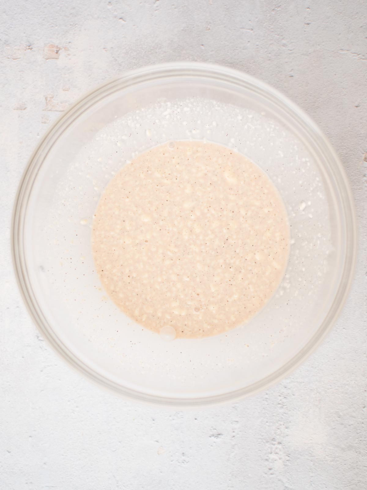 A clear glass bowl containing a bubbly, creamy beige sourdough starter on a light-colored surface. The texture is slightly thick with small air bubbles visible throughout.