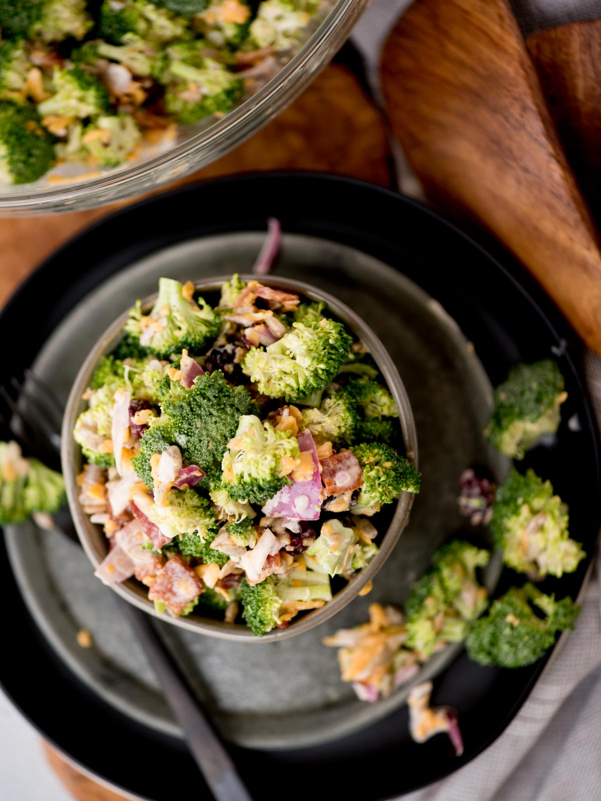 A bowl of fresh broccoli salad with chopped onions, bacon bits, and shredded cheese, placed on a black plate. A fork rests beside the bowl, and a wooden board is partially visible in the background.