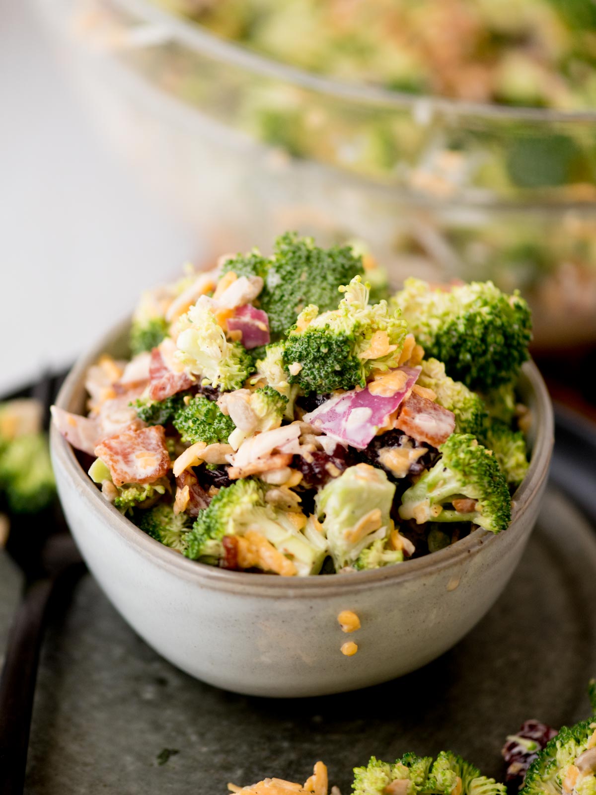 A small bowl filled with a colorful broccoli salad, featuring broccoli florets, diced red onion, bacon bits, shredded cheese, and a creamy dressing. The bowl is placed on a dark tray, with a larger bowl of salad blurred in the background.
