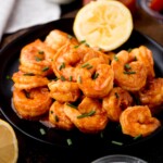 A black plate filled with seasoned, saucy buffalo shrimp garnished with chopped chives. A lemon slice graces the plates edge, while the background reveals hints of more lemon slices nearby.