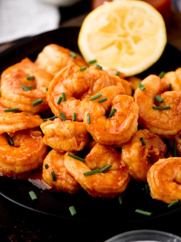 A black plate filled with seasoned, saucy buffalo shrimp garnished with chopped chives. A lemon slice graces the plates edge, while the background reveals hints of more lemon slices nearby.