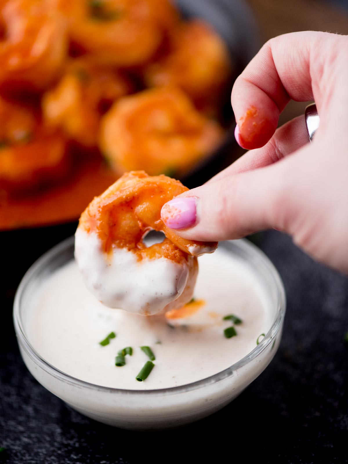 A hand with colorful nails dips a breaded and saucy shrimp into a bowl of creamy white dip, garnished with chopped chives. A bowl of coated shrimp is visible in the background.