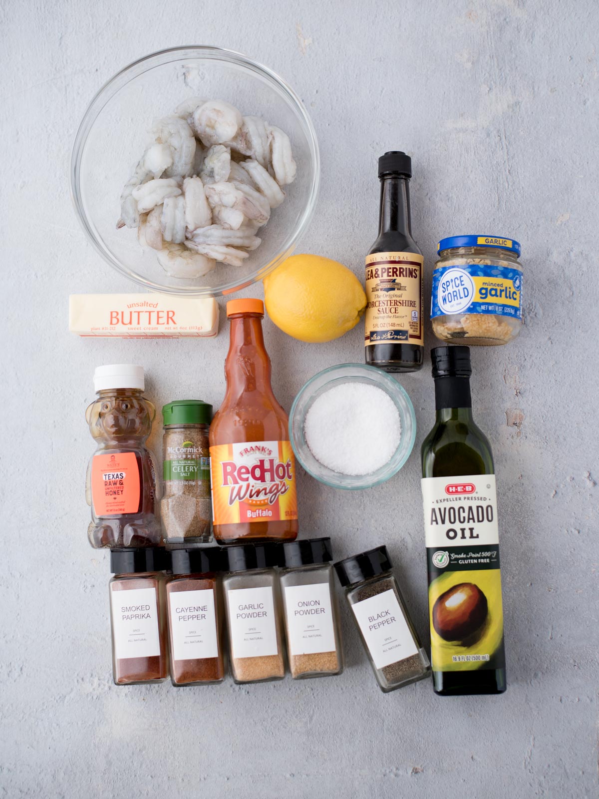A flat lay of cooking ingredients on a light surface: raw shrimp in a bowl, a stick of butter, Worcestershire sauce, minced garlic, lemon, hot sauce, salt, and various spices. Theres also a jar of avocado oil.