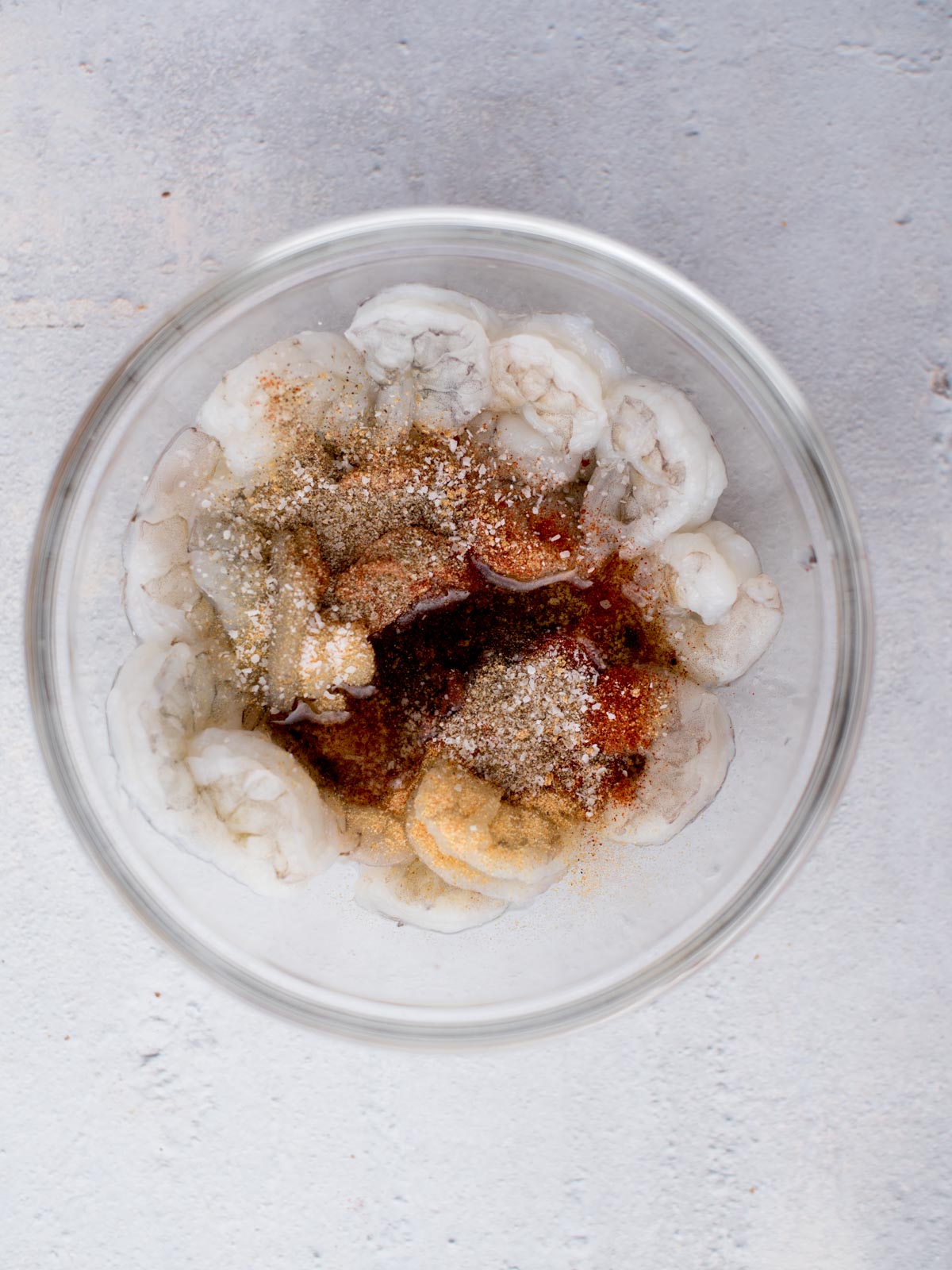 A glass bowl filled with raw shrimp seasoned with various spices. The shrimp are arranged in a circular pattern against a light gray background.