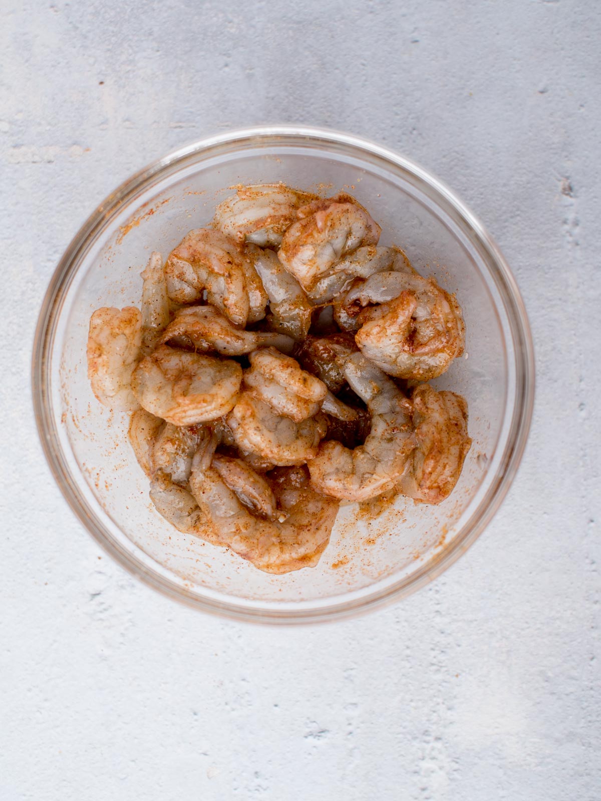 A glass bowl filled with raw, deveined shrimp seasoned with spices, placed on a light gray surface.