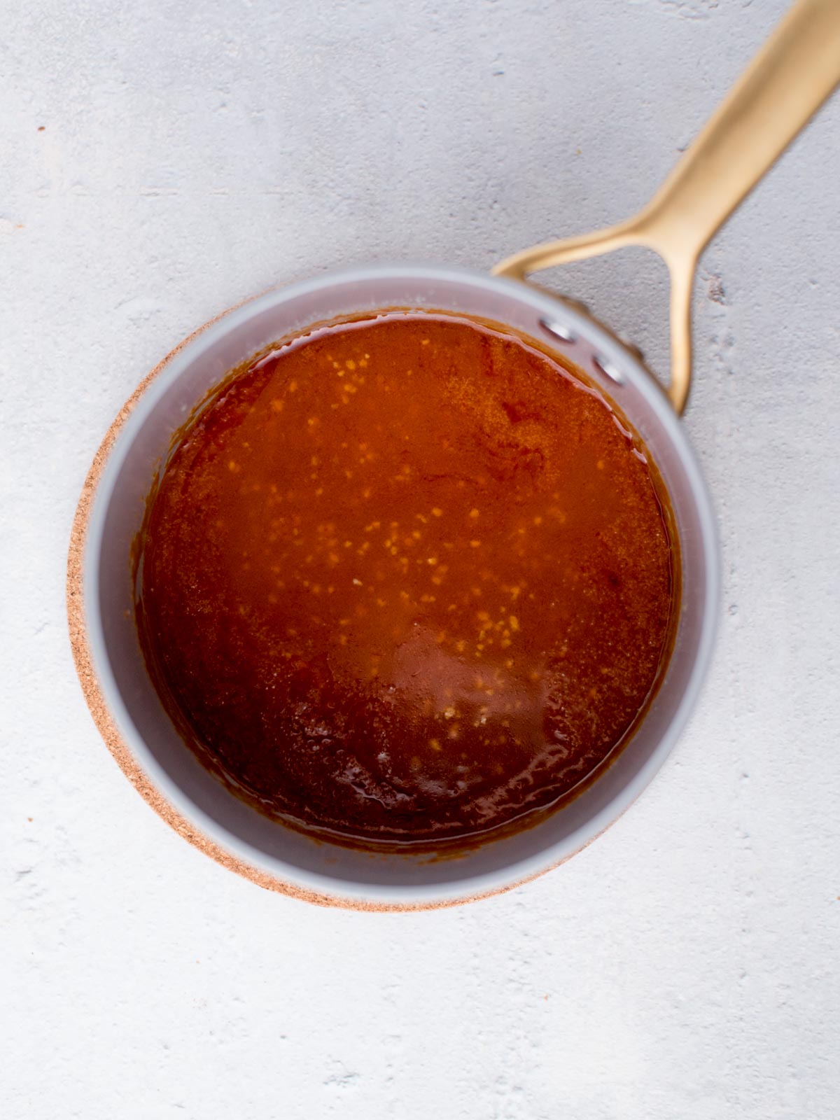 Top view of a saucepan with golden buffalo sauce against a light background. The sauce appears smooth and glossy, with a few bubbles around the edges. The saucepan has a golden handle.