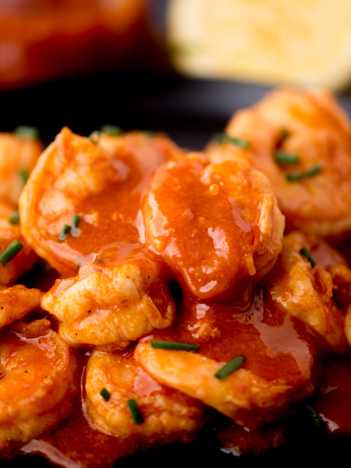Close-up of shrimp covered in a spicy red sauce, garnished with finely chopped green herbs. The background features a blurred lemon wedge and a hint of a sauce container.