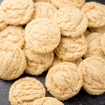A pile of round, golden brown pudding cookies with a crinkled surface, arranged closely together on a dark surface.
