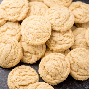 A pile of round, golden brown pudding cookies with a crinkled surface, arranged closely together on a dark surface.