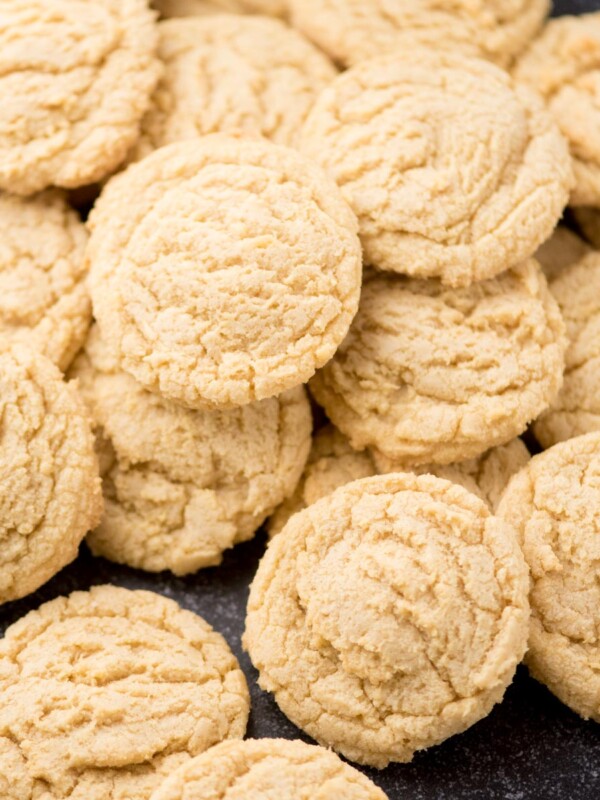 A pile of round, golden brown pudding cookies with a crinkled surface, arranged closely together on a dark surface.