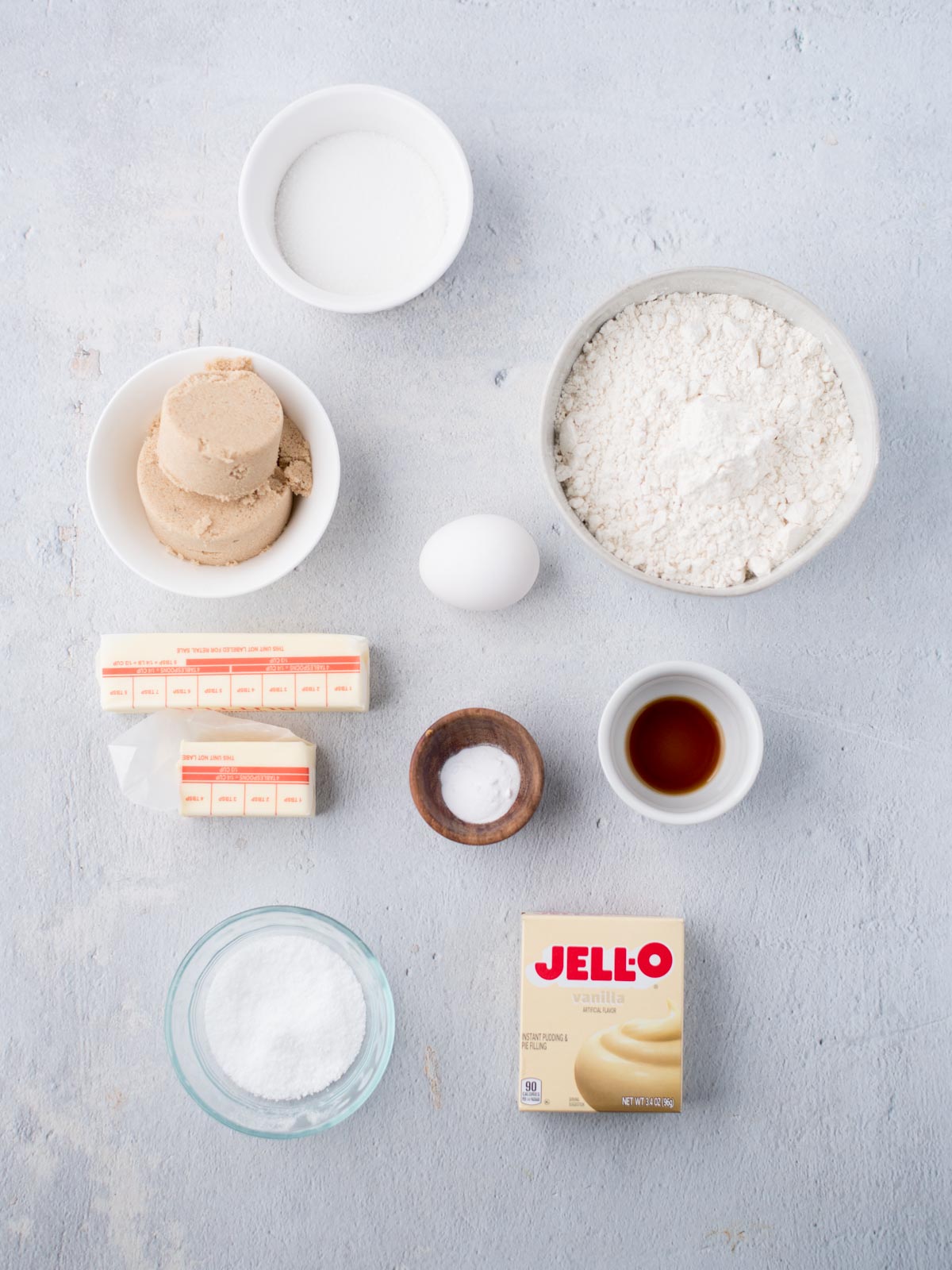 A top-down view of baking ingredients neatly arranged on a light surface, ready to make delicious vanilla pudding cookies, featuring flour, egg, sugar, brown sugar, butter, baking powder, baking soda, vanilla extract, and JELL-O pudding mix.