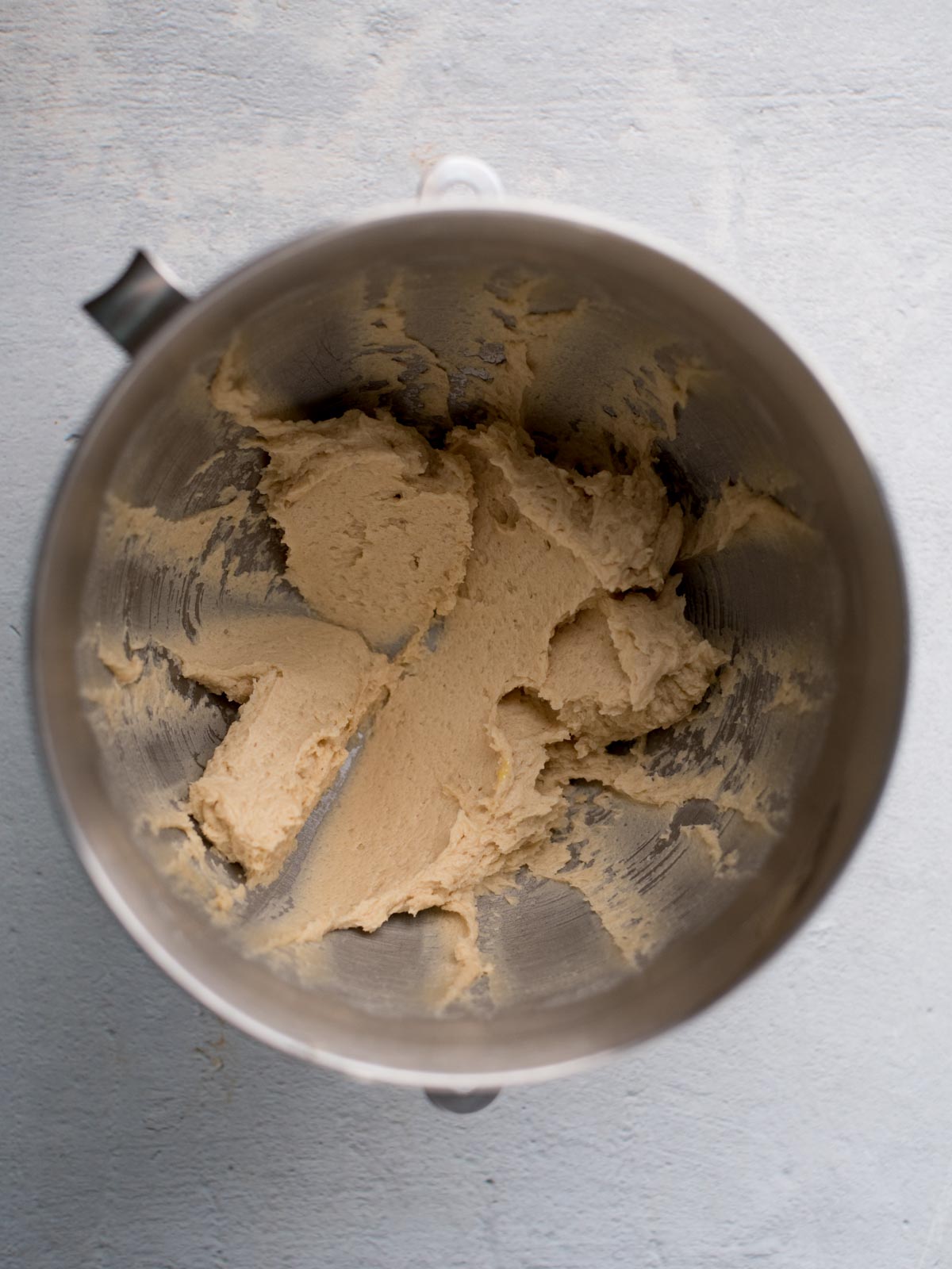 A metal mixing bowl containing creamed pudding cookies dough, with some dough stuck to the sides of the bowl. The surface below the bowl is light gray.
