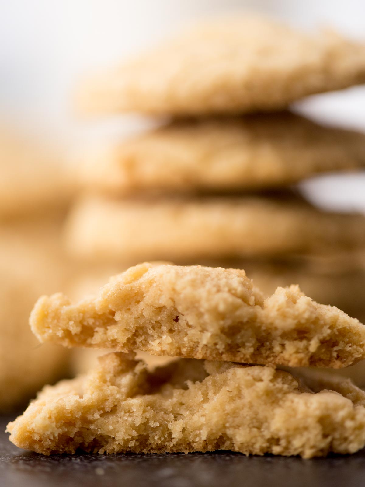 A close-up of a stack of crumbly, golden brown pudding mix cookies, with one cookie broken in half in the foreground, showing its soft, textured inside. The background is softly blurred.