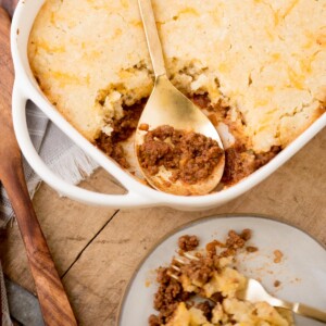 A white baking dish filled with a savory pie topped with golden, crumbly crust. A serving is missing, revealing a minced meat filling. A gold spoon rests inside. Nearby, a plate holds a portion with meat and crust. A wooden board and utensils are visible.