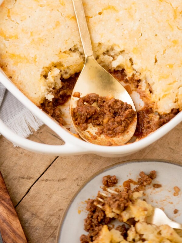 A white baking dish filled with a savory pie topped with golden, crumbly crust. A serving is missing, revealing a minced meat filling. A gold spoon rests inside. Nearby, a plate holds a portion with meat and crust. A wooden board and utensils are visible.