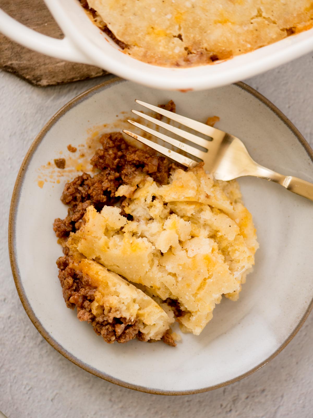 A plate with a serving of shepherds pie, featuring a layer of golden-brown mashed potatoes over seasoned ground meat. A fork rests on the plate. Part of the baking dish is visible in the top left corner.