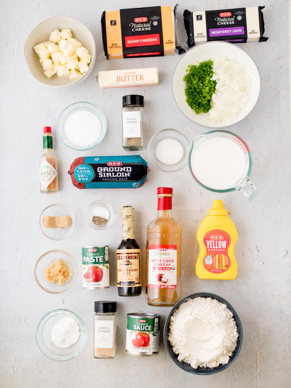 Ingredients laid out on a gray surface include cheese blocks, butter, minced garlic, chopped onions, milk, ground beef, hot sauce, Worcestershire sauce, ketchup, mustard, flour, cornstarch, jalapeños, salt, pepper, and spices.