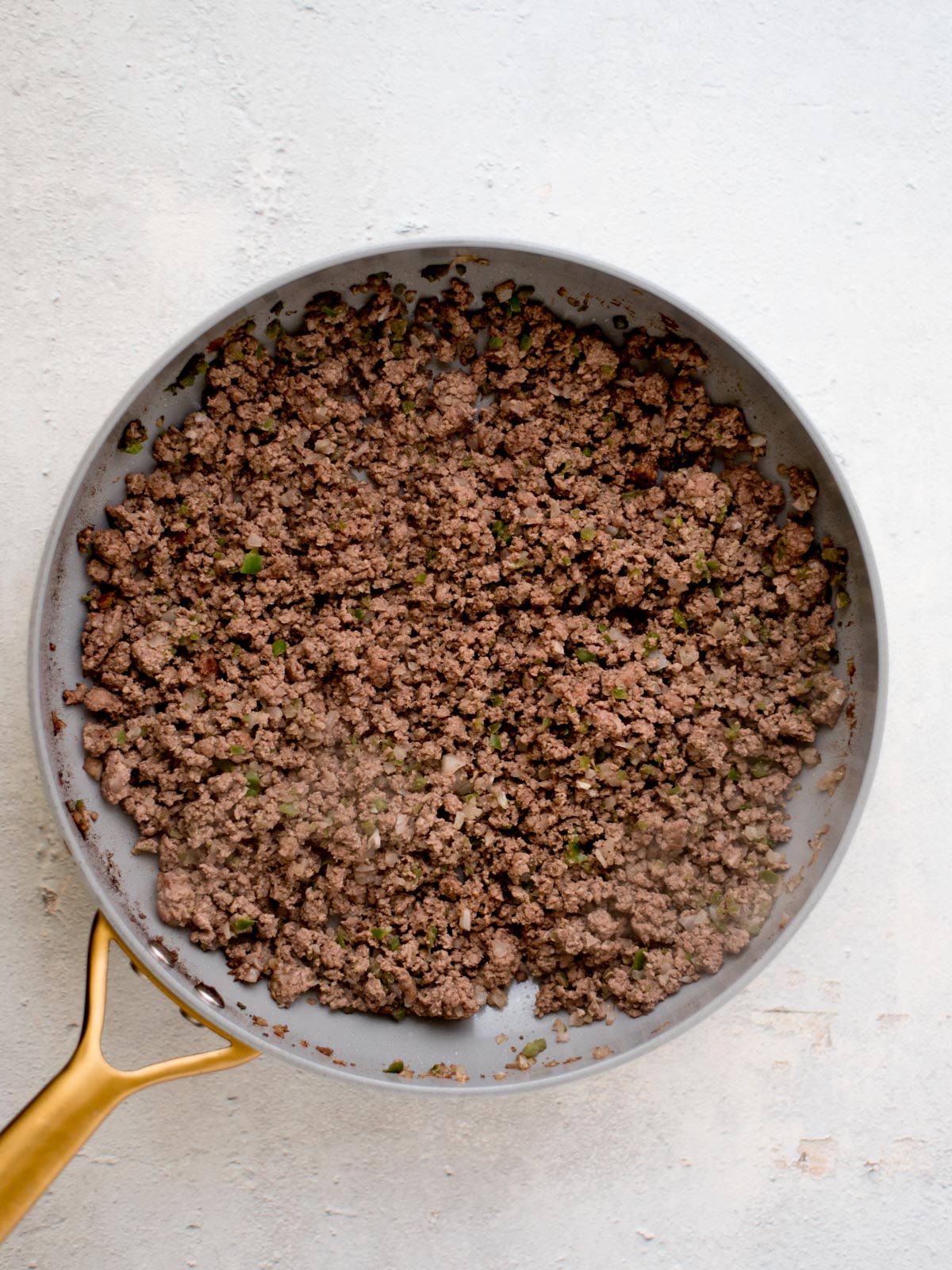 A frying pan with a gold handle is filled with browned ground beef on a light countertop. Some herbs are mixed into the meat, adding specks of green. The surface of the meat is slightly textured and crumbly.