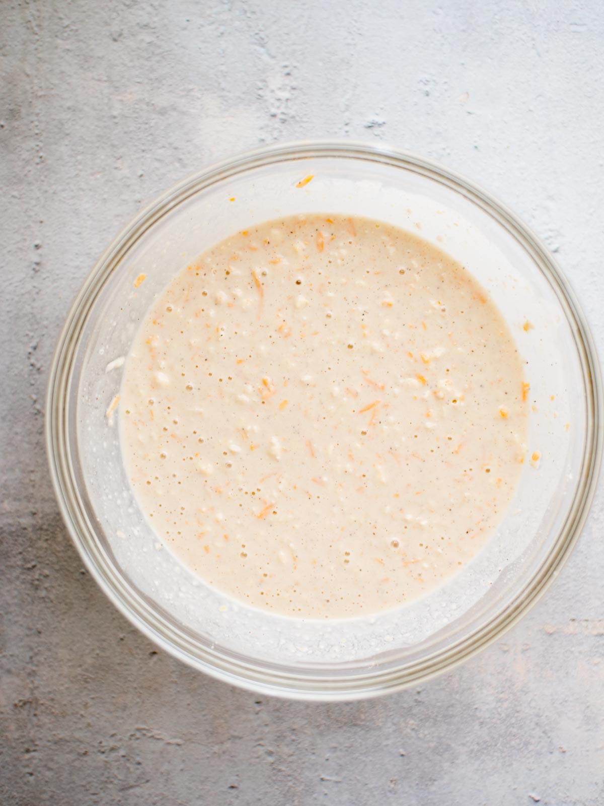 A glass bowl filled with a light beige batter containing visible shreds of orange carrots, sitting on a textured gray surface.