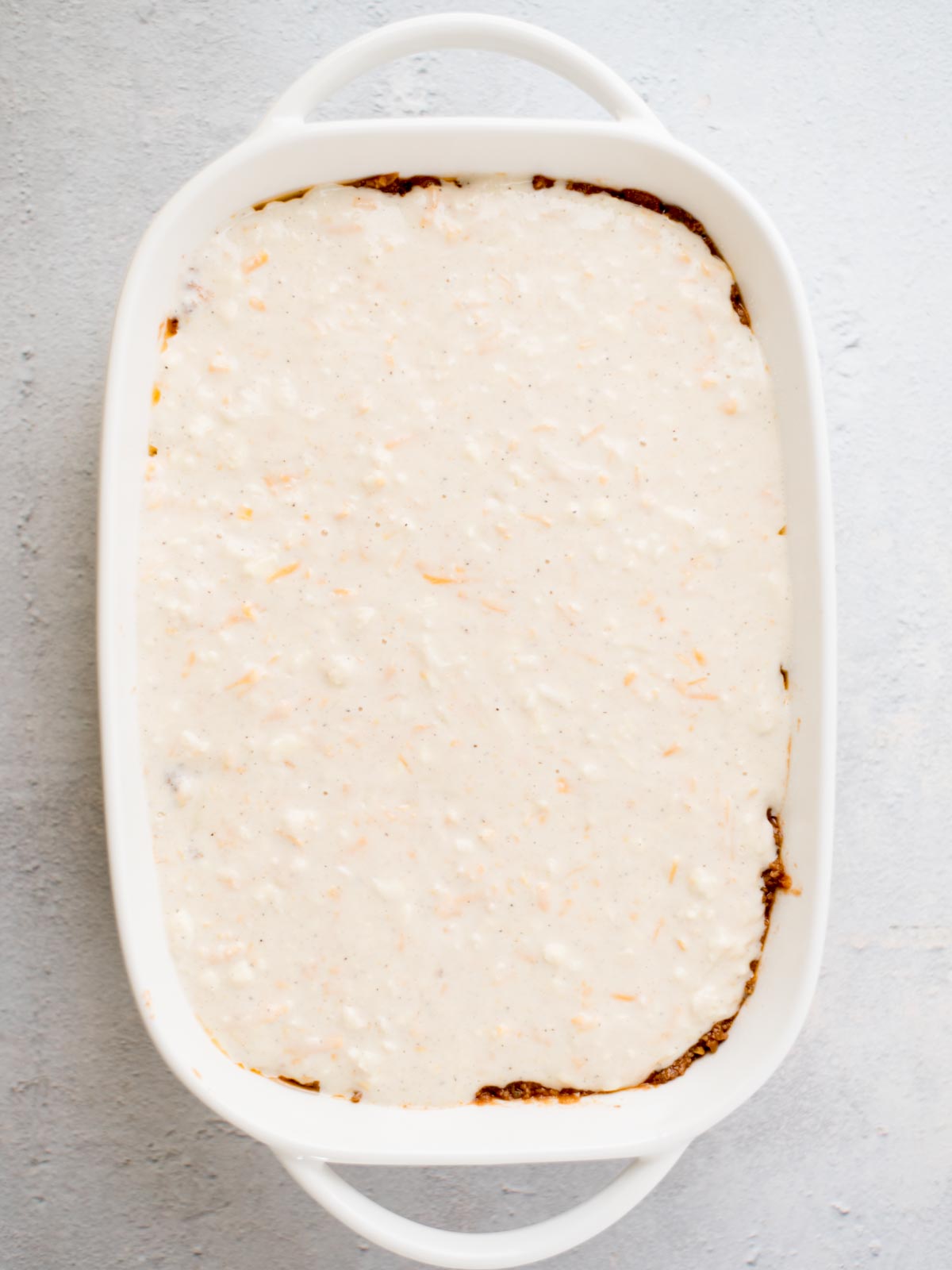 A rectangular white casserole dish filled with a layered dish. The top layer is a creamy white sauce with grated cheese visible throughout. The dish is placed on a light gray surface.