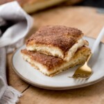 Two square pieces of cinnamon sugar cheesecake bars stacked on a white plate. Next to them is a gold fork. The bars have a flaky crust and are topped with a cinnamon-sugar mixture. A cloth napkin is partially visible to the side.