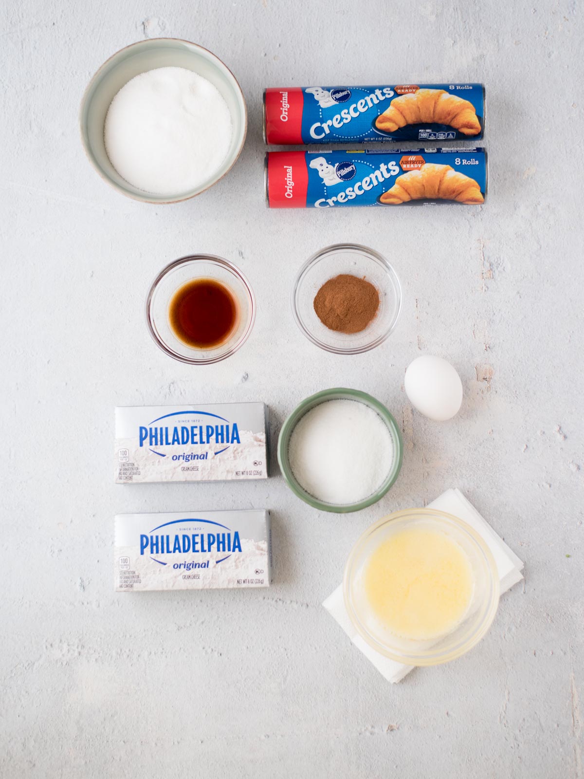 Ingredients for a crescent roll cheesecake recipe are arranged on a light surface: two crescent roll packages, a bowl of sugar, vanilla extract, ground cinnamon, cream cheese, an egg, melted butter, and a bowl of sugar.