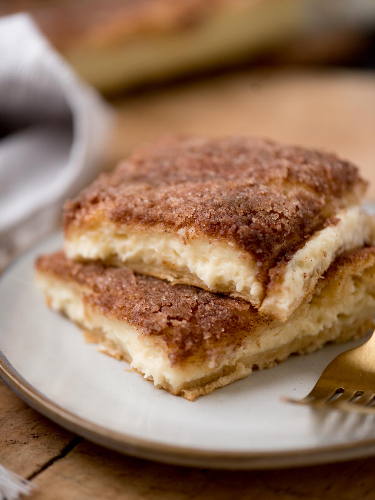 Two slices of cinnamon sugar sopapilla cheesecake on a plate, revealing a creamy filling. A golden fork rests beside them. The background is softly blurred, highlighting the desserts flaky texture and sugary crust.