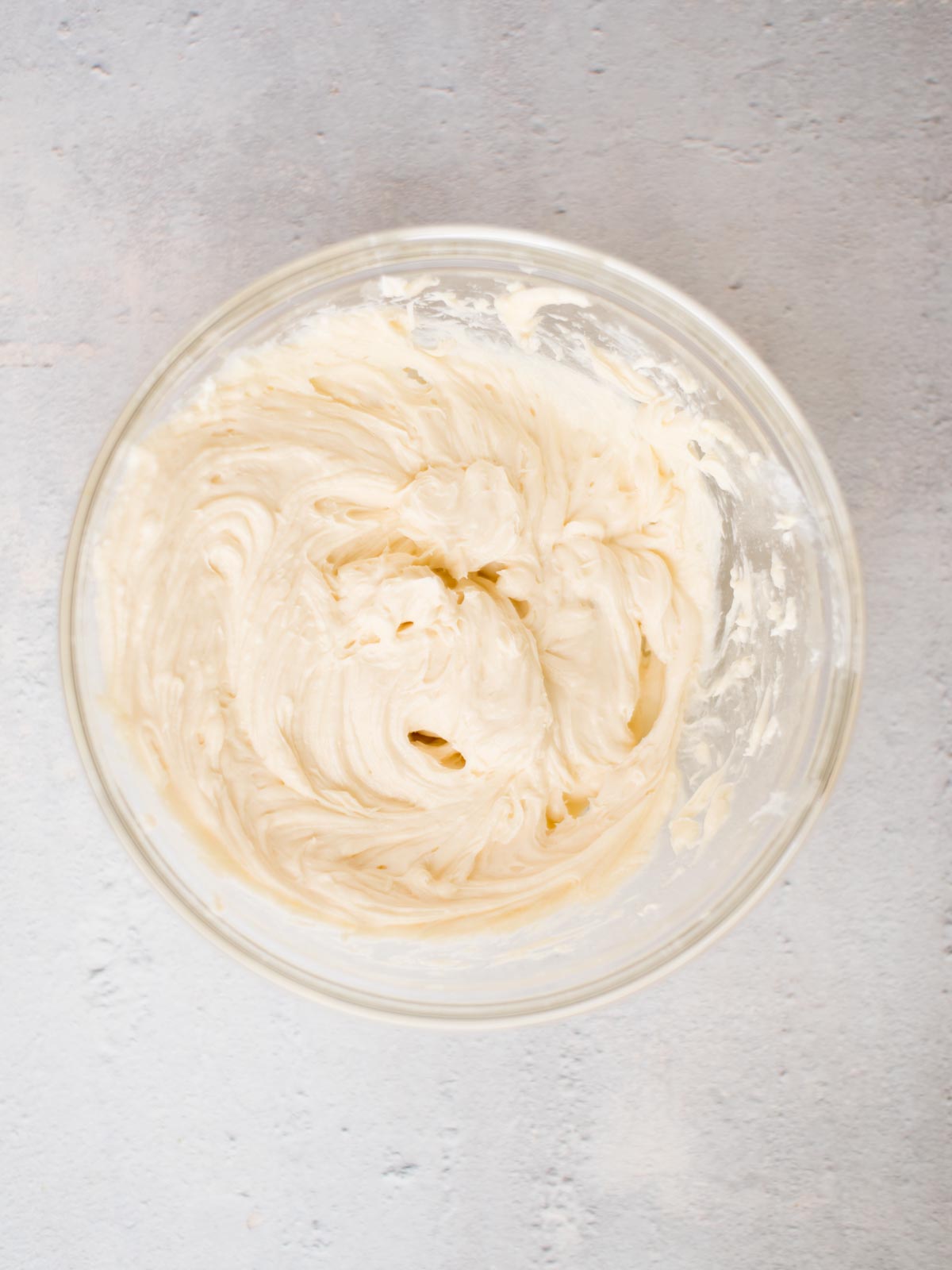 A glass bowl filled with creamy, whipped vanilla frosting sits on a light gray textured surface. The frosting is smooth and swirled, indicating it has been freshly mixed.